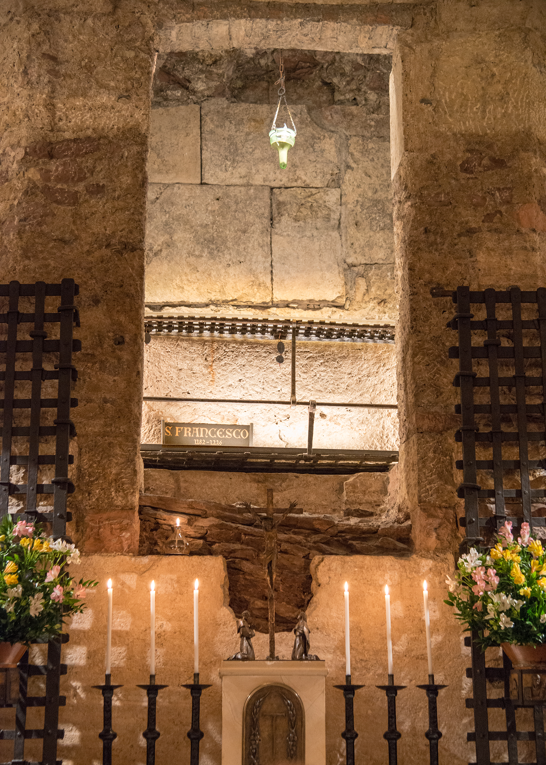The Tomb of Saint Francis underneath the basilica is inside a crypt chapel excavated from the distinctive pink rock of the mountains outside Assisi. A pillar of stone rises to the center of a dome; carved inside is the tomb of Saint Francis of Assisi. In front of the pillar is a small altar with simple candlesticks and a tabernacle depicting Saint Francis and Saint Clare kneeling before the Cross. Candles fill the tomb with peaceful, warm light.